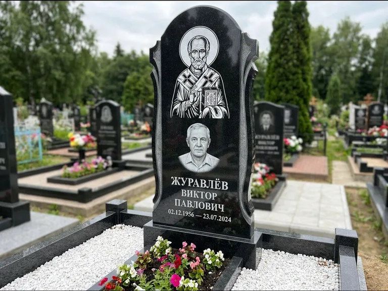 Памятник с гравировкой иконы: стела с образом в полный рост на обороте — гравировка образа Матроны Московской, низкие кусты спиреи в цвету, работа «Лебедева» в Санкт-Петербурге